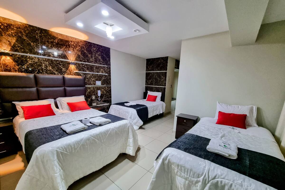 A hotel room with three beds, each with white bedding and red pillows, a dark headboard, and a marble accent wall.