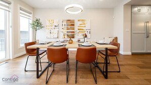 Een moderne eetkamer met een lange tafel, zes stoelen en een kroonluchter. Aan de muur hangen abstracte schilderijen en er staat een koelkast op de achtergrond.