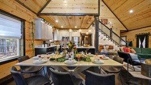 A spacious dining area with a long table set for a meal, surrounded by modern chairs. A staircase leads to an upper level, and the room features wooden walls and ceilings.
