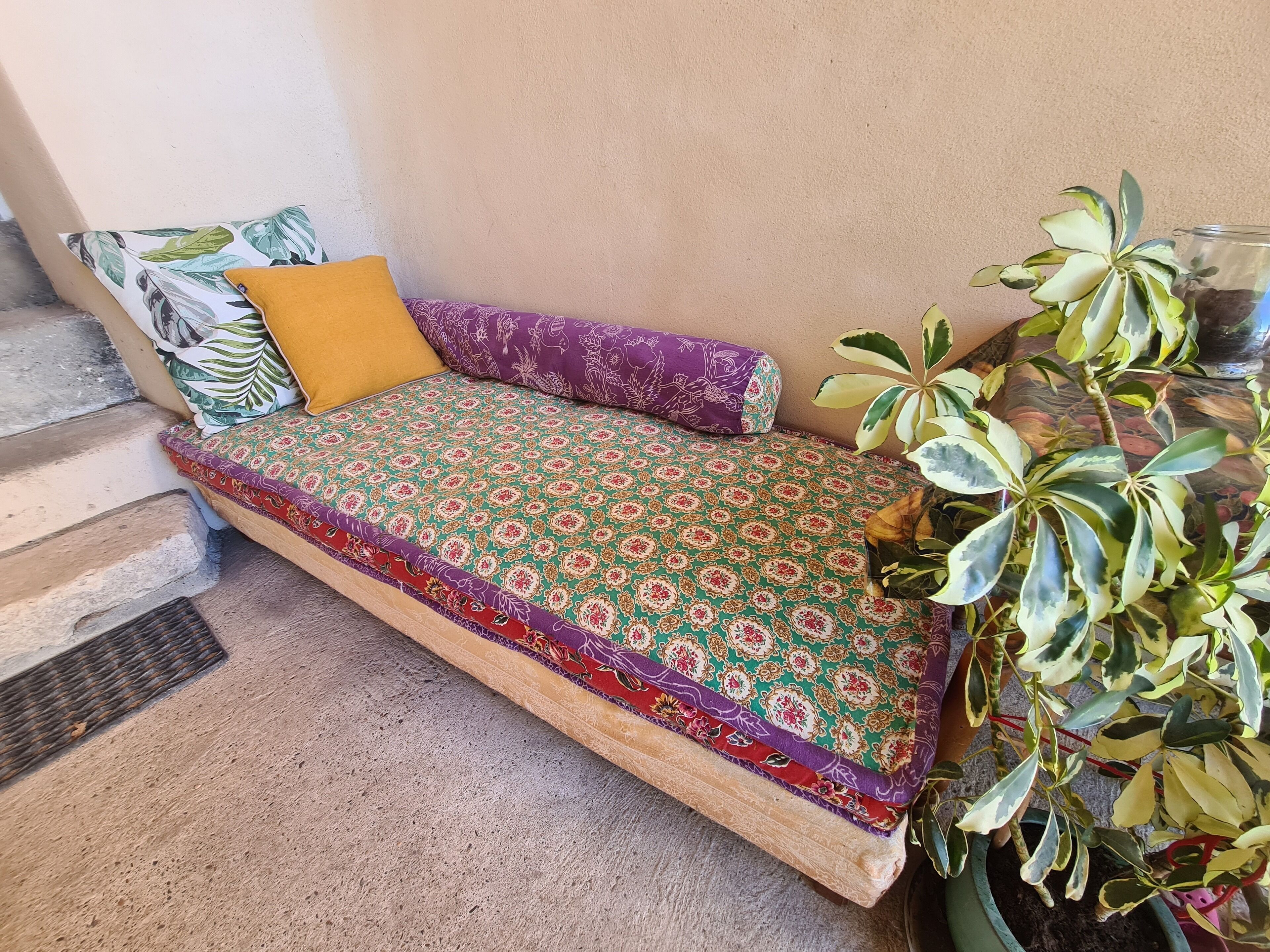 A small, cozy bed with patterned bedding, a yellow pillow, and a purple pillow against a plain wall.
