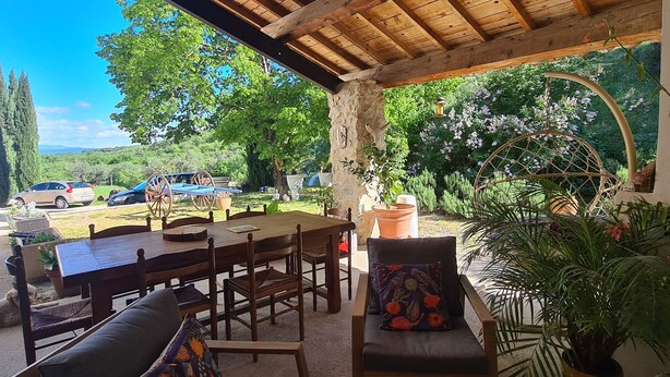 Une terrasse extérieure avec des meubles en bois, un mur en pierre et une vue sur les voitures garées et la verdure.