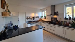 Une cuisine moderne avec des armoires blanches, des comptoirs noirs et un îlot central. On y trouve des casseroles et des poêles suspendues, un four encastré et une fenêtre donnant sur l’extérieur.