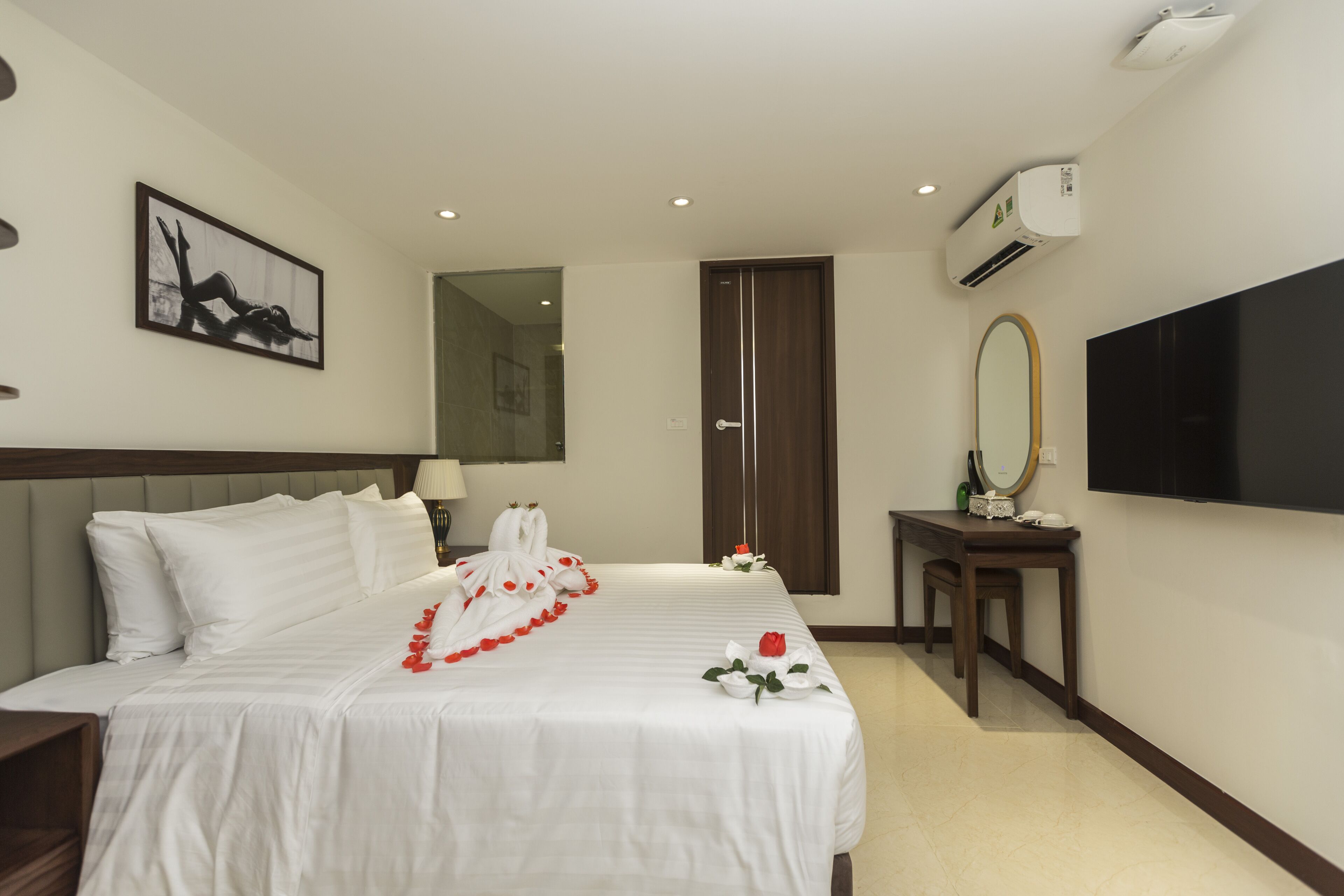 A hotel room with a neatly made bed, a bedside table with a lamp, a mirror, and a flat-screen TV mounted on the wall.