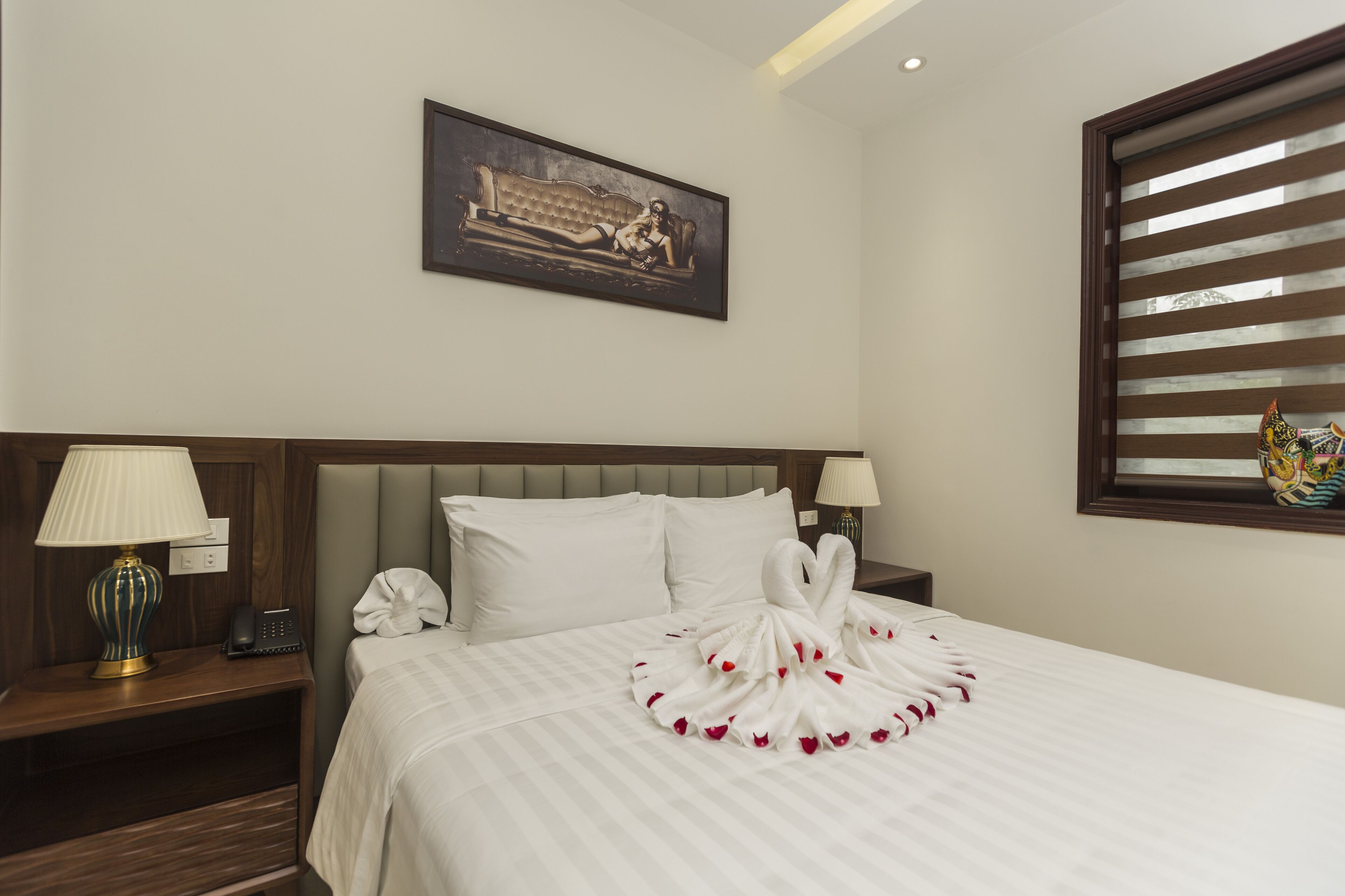 A neatly made bed with a swan towel centerpiece, a bedside lamp, a phone, and a painting on the wall.