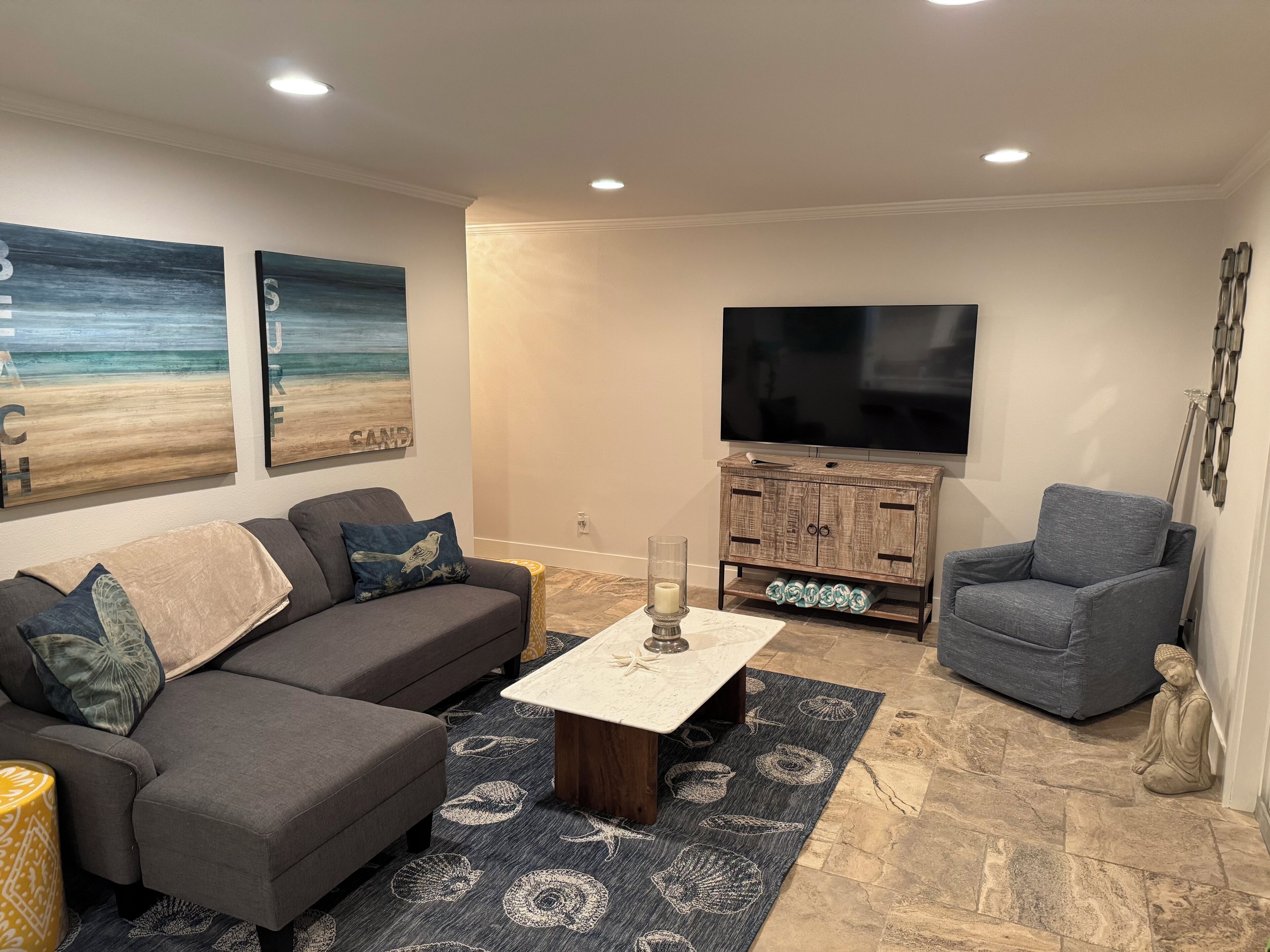A living room with a gray sofa, a blue armchair, a coffee table, and a TV on the wall.