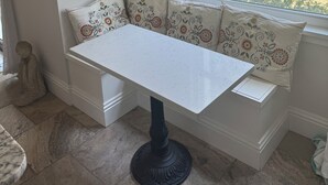 Un coin salon avec une table et un banc blancs, des éléments décoratifs muraux et une vue sur l’extérieur.