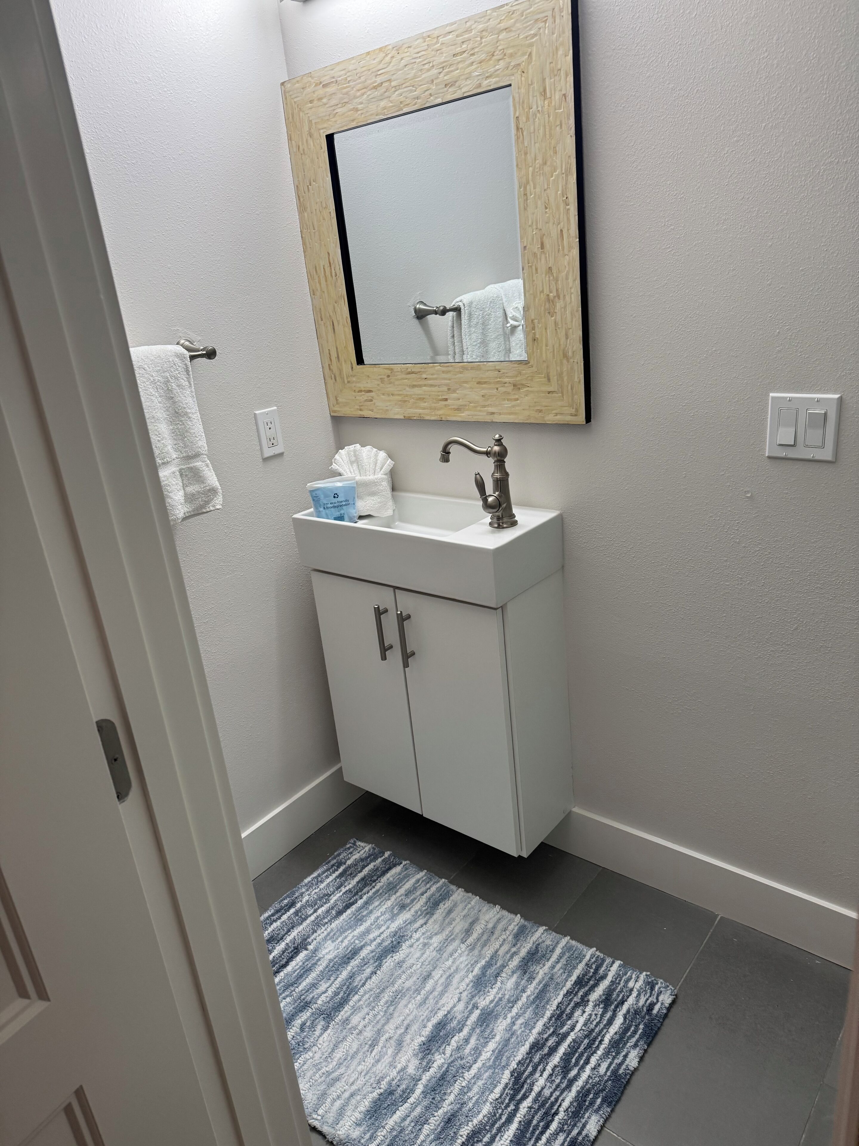 Une salle de bain avec un lavabo blanc, un miroir avec un cadre en bois, un porte-serviettes et un tapis à rayures bleues.