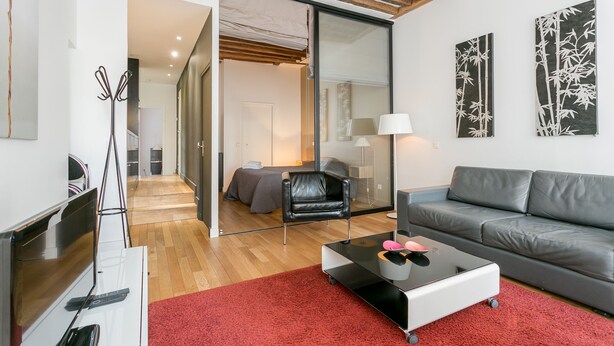 Una sala moderna con un sofá, una mesita de centro y un televisor. Hay una puerta corrediza que da a un dormitorio.