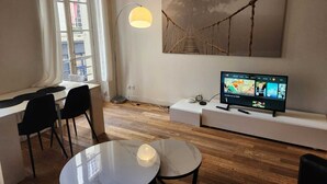 Una sala de estar moderna con piso de madera, una mesa redonda, dos sillas negras, un televisor sobre un mueble blanco, una lámpara de pie y un cuadro abstracto grande en la pared.