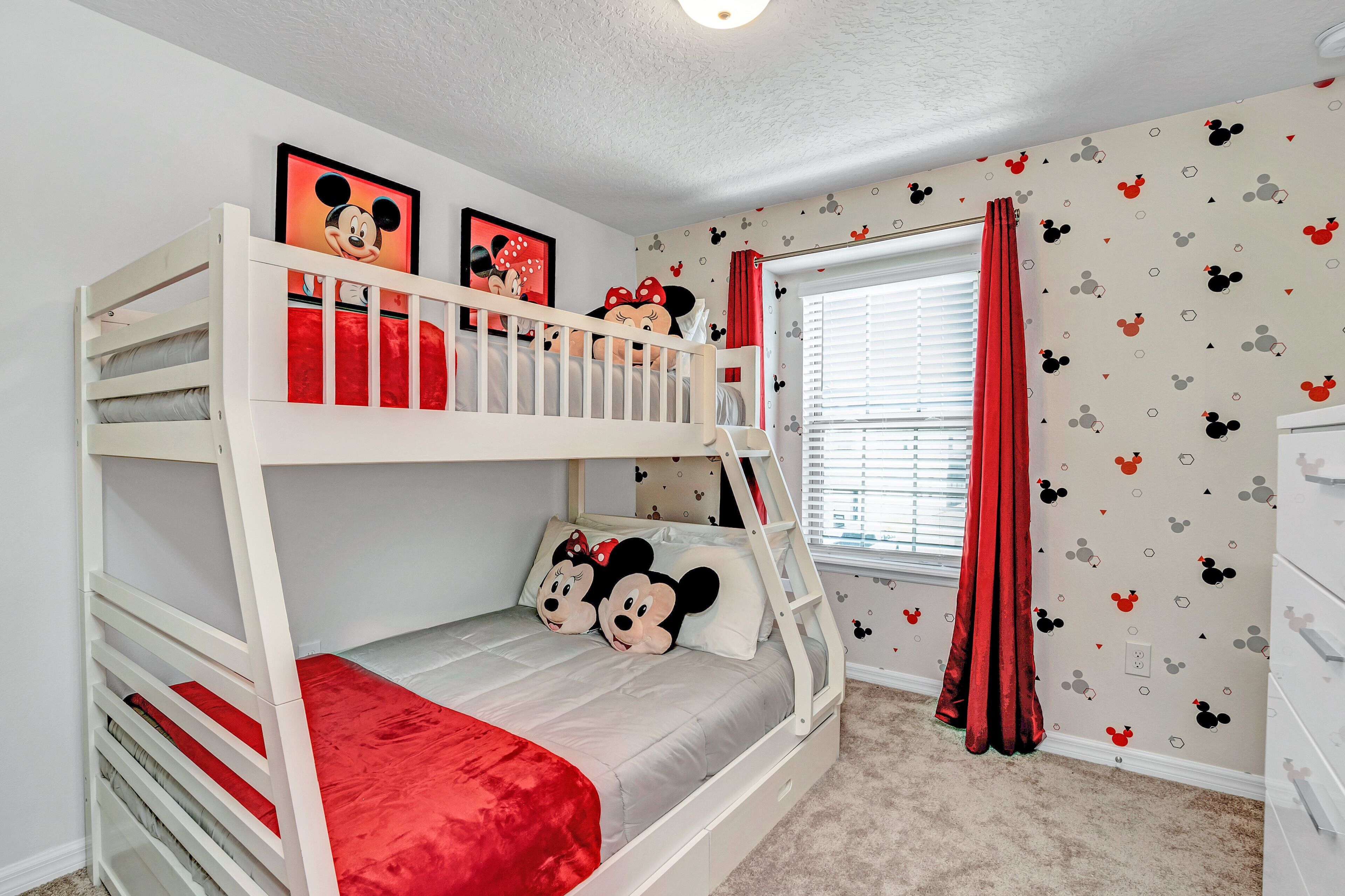 Ein Schlafzimmer mit Etagenbetten, Deko im Mickey-Mouse-Stil, einem Fenster mit Jalousien und einem roten Vorhang.