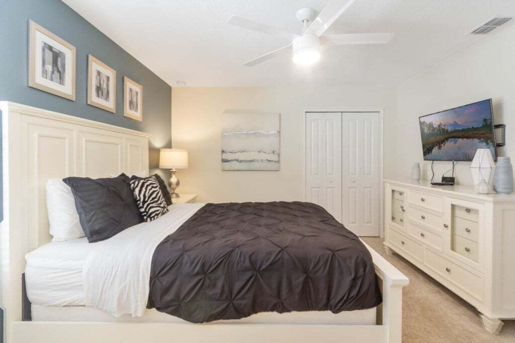 A bedroom with a bed, a nightstand, a dresser, a ceiling fan, and a painting on the wall.