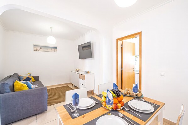 A modern living room with a sofa, dining table set for two, and a TV mounted on the wall.
