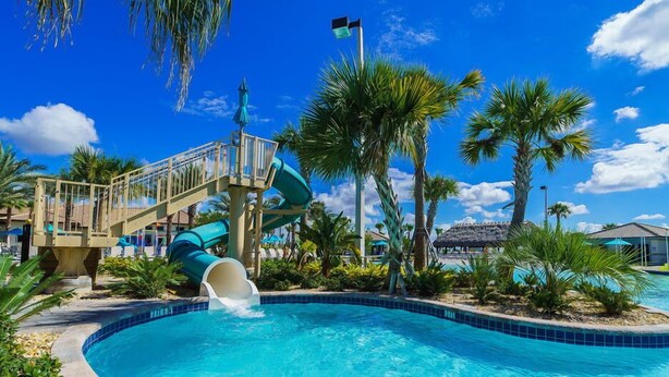 A pool with a water slide, palm trees, and a thatched-roof structure.