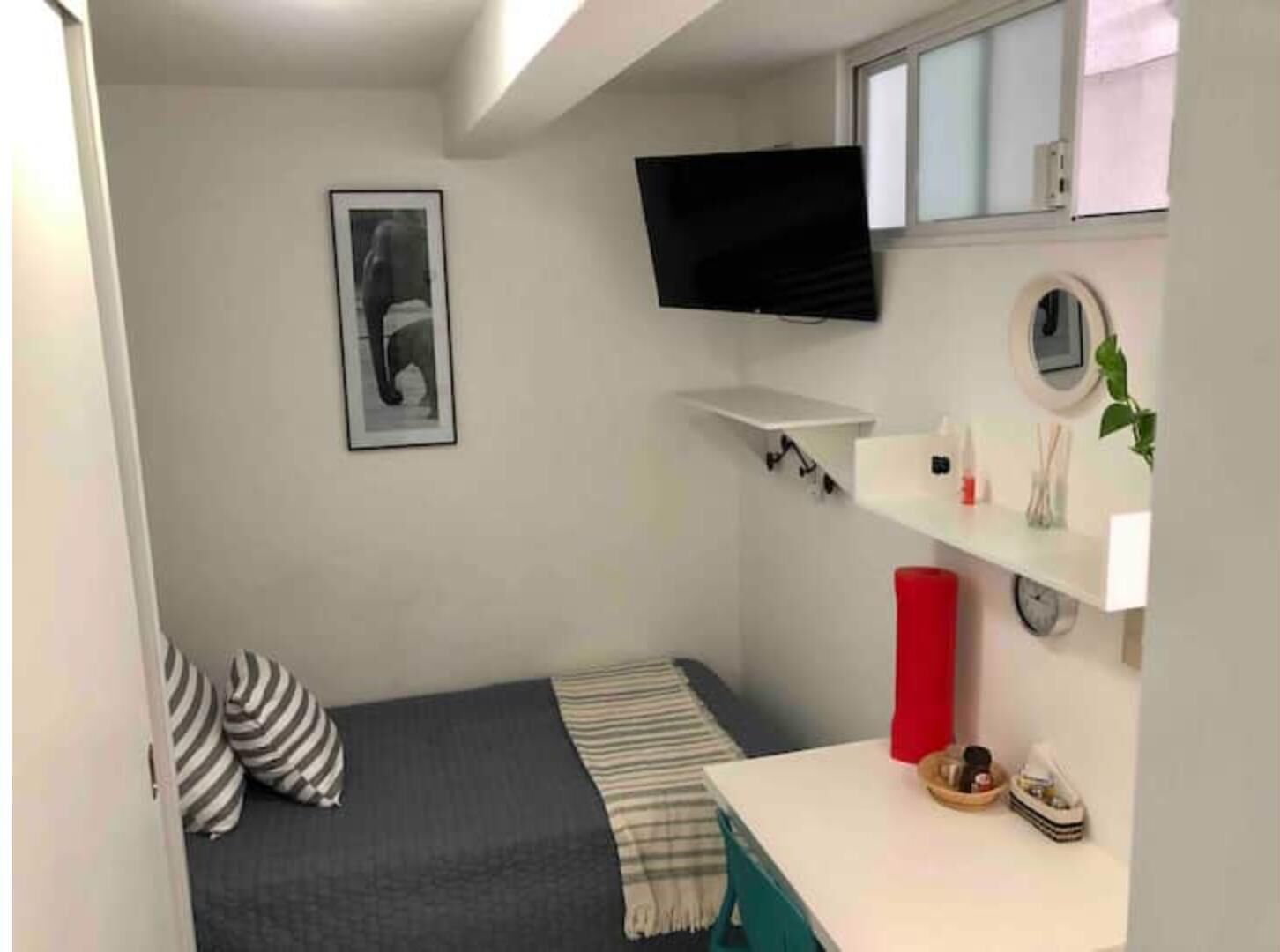 Ein kleiner, kompakter Raum mit einem Bett, einem an der Wand montierten Fernseher, einer kleinen Ablage mit Pflegeprodukten und einem Fenster.