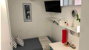 A small, compact room with a bed, a TV mounted on the wall, a small shelf with toiletries, and a window. - Apartaestudio#4 Itesm Pequeño, Acogedor & Céntrico (Santiago de Querétaro)