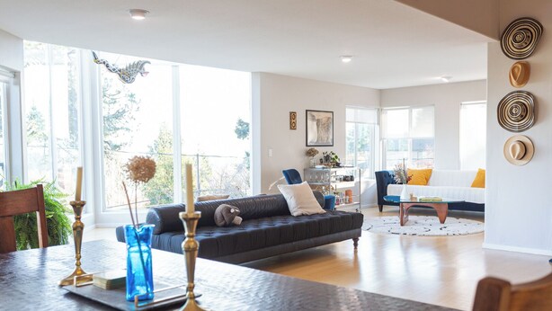 A spacious living room with a dark grey sofa, a coffee table, and a dining area with a wooden table and blue glassware. - PDX Penthouse with Mountain & River Views (Portland)
