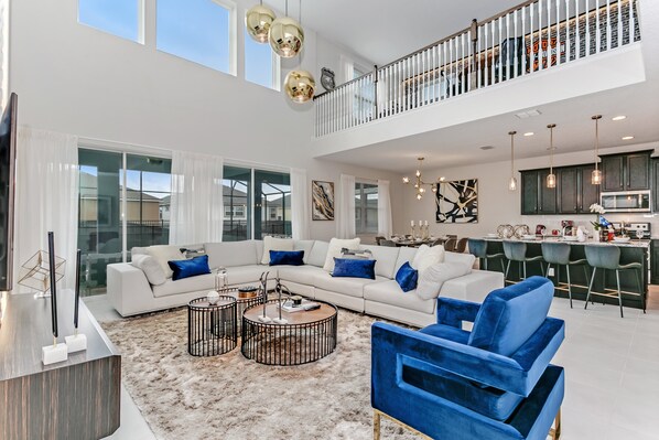 A spacious living area with a sectional sofa, a coffee table, and a blue armchair. There is a kitchen in the background with bar stools.