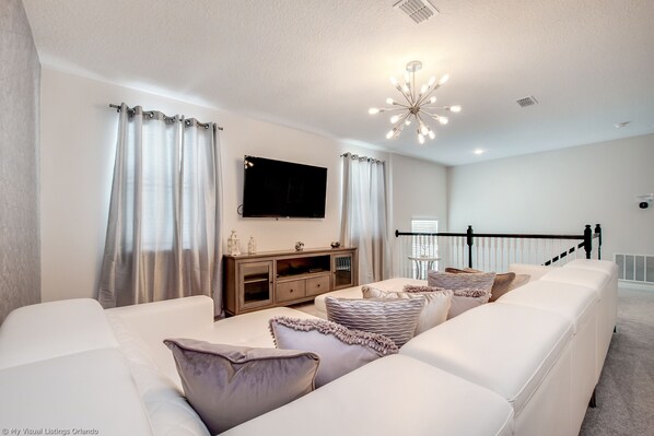 A modern living room with a large white sofa, a wooden entertainment unit, and a flat-screen TV mounted on the wall.