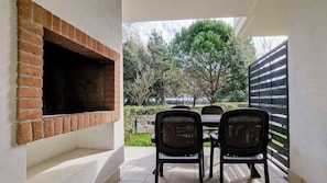 Une cheminée en briques, un espace extérieur aménagé avec des chaises, et une vue sur les arbres et une clôture.