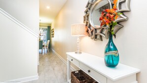 A hallway with a white console table, a teal vase with flowers, a patterned lamp, and a decorative mirror.