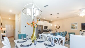 A modern dining room with a white table set for four, a decorative vase, and a kitchen in the background.
