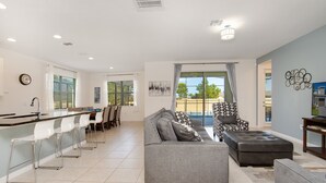 A modern living room with a grey sofa, ottoman, and a patterned armchair. There is a kitchen area with white bar stools and a dining table. Large windows provide natural light and a view of the outdoors.