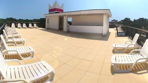 A rooftop terrace with white lounge chairs arranged in rows, a small building with a crown-like structure, and a clear sky.