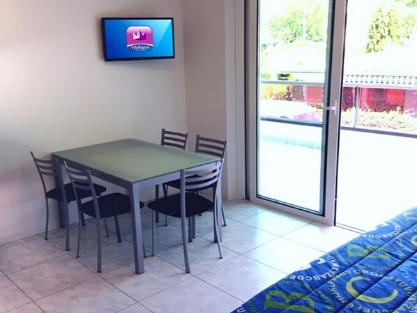 A room with a dining table and chairs, a TV mounted on the wall, and a large window with a view of the outside.