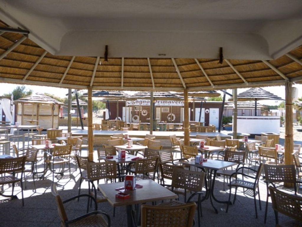 Un café en plein air, avec des tables et des chaises sous un toit de chaume, entouré de bâtiments.