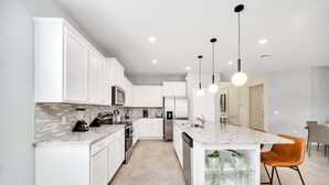 A modern kitchen with white cabinets, granite countertops, and stainless steel appliances. There are pendant lights above the island and a plant on the lower shelf. - Last Minute Disney Getaway! Private Pool, Hot Tub & Game Room. Book Now! #42814 (Orlando)