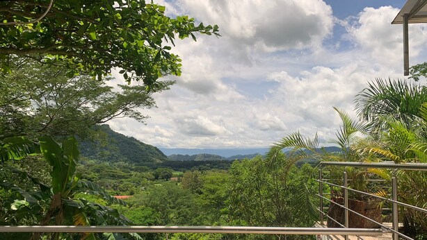 A balcony with a metal railing overlooking a lush green landscape with hills and a partly cloudy sky.