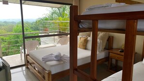 A room with a wooden bunk bed, a small bedside table, a chair, and a large sliding glass door leading to a balcony with a view of greenery.