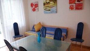A room with a glass dining table, blue chairs, a sofa with a yellow pillow, and artwork on the walls.