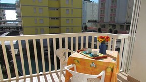 A balcony with a table set for two, a vase with flowers, and a view of surrounding buildings.
