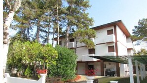 A multi-story building with a white facade and brown accents, surrounded by trees and a fenced garden area. - Residenz Codan Wohnung Codan für 6 Personen (Bibione)