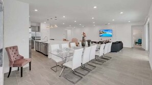 A modern kitchen with white cabinets, a dining area with a glass table and white chairs, and a TV mounted on the wall.
