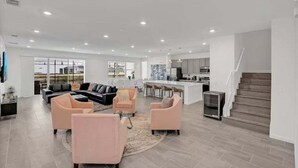 A spacious living area with a black sectional sofa, two pink armchairs, and a kitchen area with a bar counter and modern appliances.