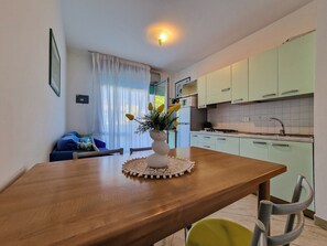 A dining table with a decorative centerpiece, a kitchen with light green cabinets, and a window with sheer curtains. - Residenz Argonauti Wohnung Trilo C2 1S für 6 Personen (Bibione)