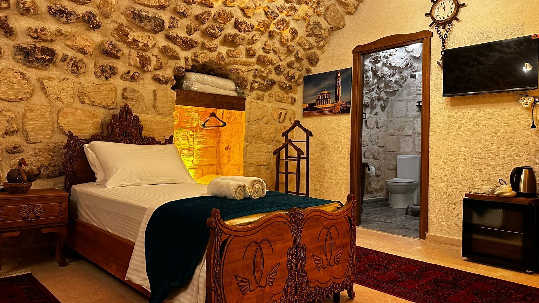 A bedroom with a stone wall, a bed with a wooden headboard, a wooden nightstand, a wall-mounted TV, and a bathroom visible through an open door.