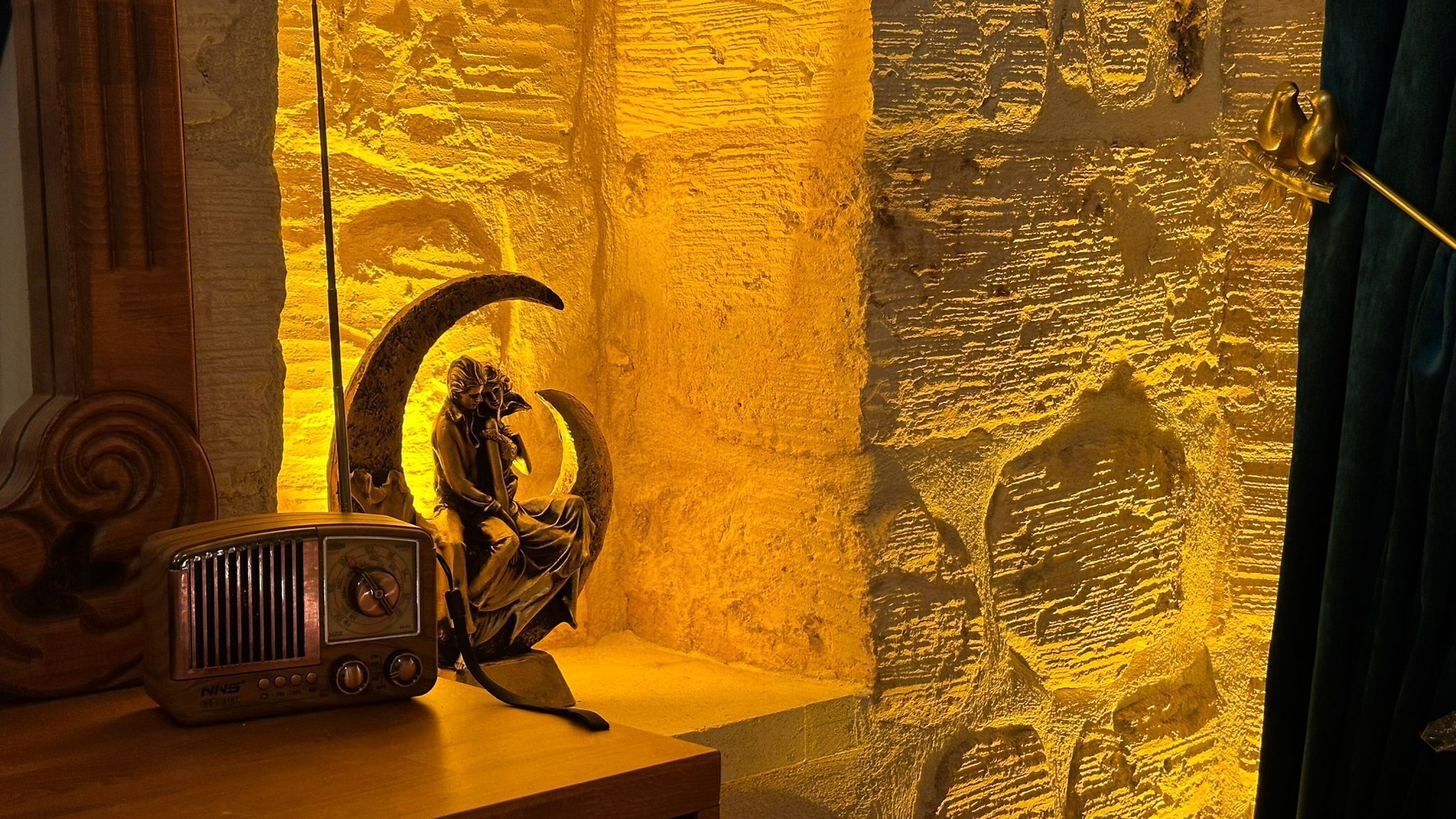 Una radio antigua sobre una mesa de madera, una escultura de una mujer y una luna creciente, y una pared texturizada con una puerta y una cortina.