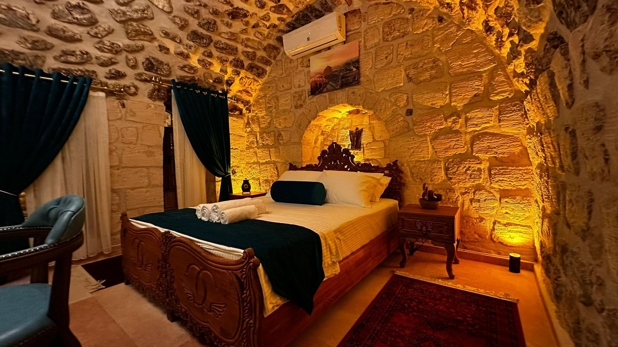Un dormitorio con pared de piedra, una cama de madera, una mesita de noche, una silla y una alfombra roja.