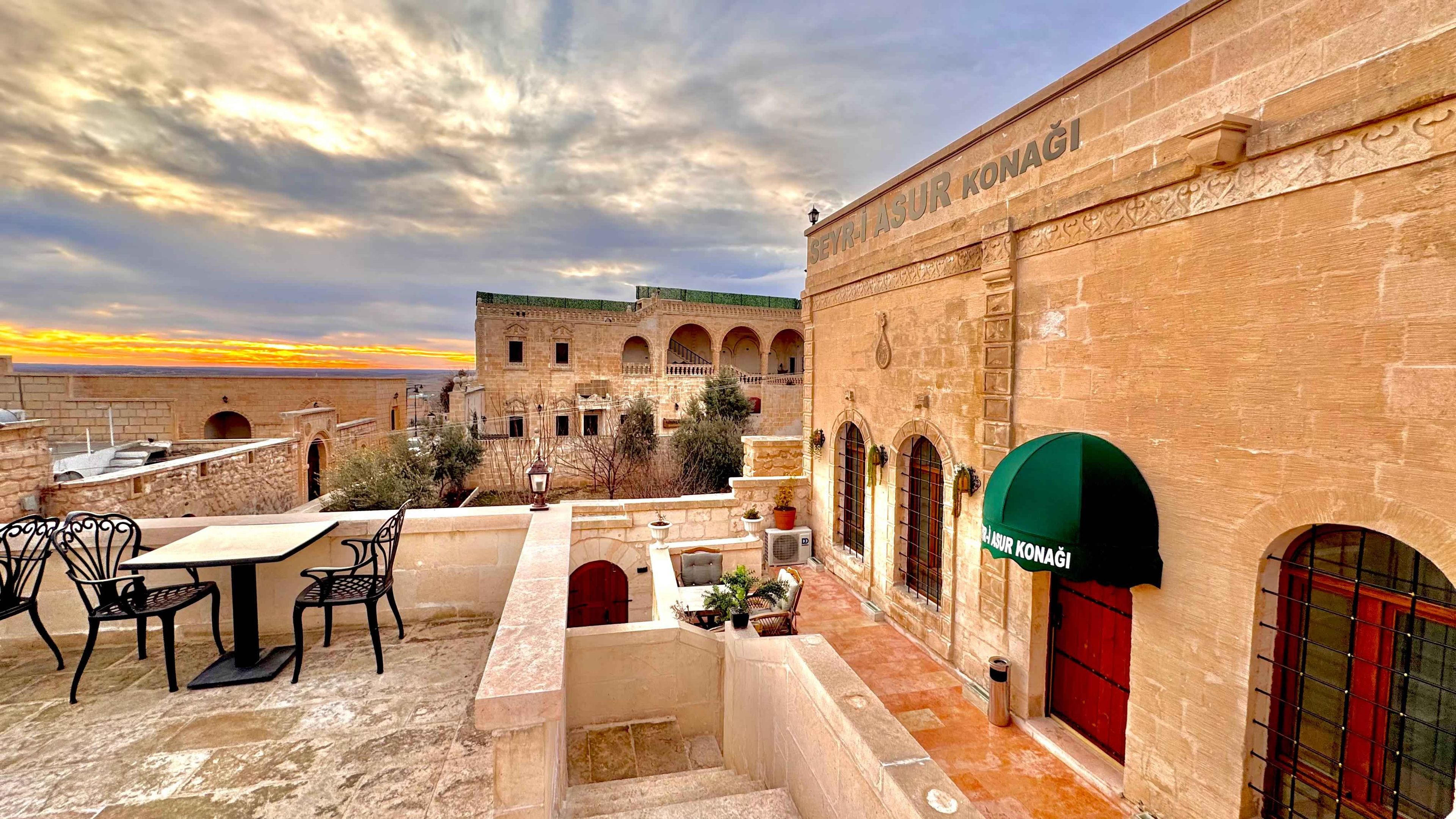 Un patio con asientos al aire libre, un edificio con un letrero que dice 'SEYRI ASUR KONAGI' y un atardecer al fondo.