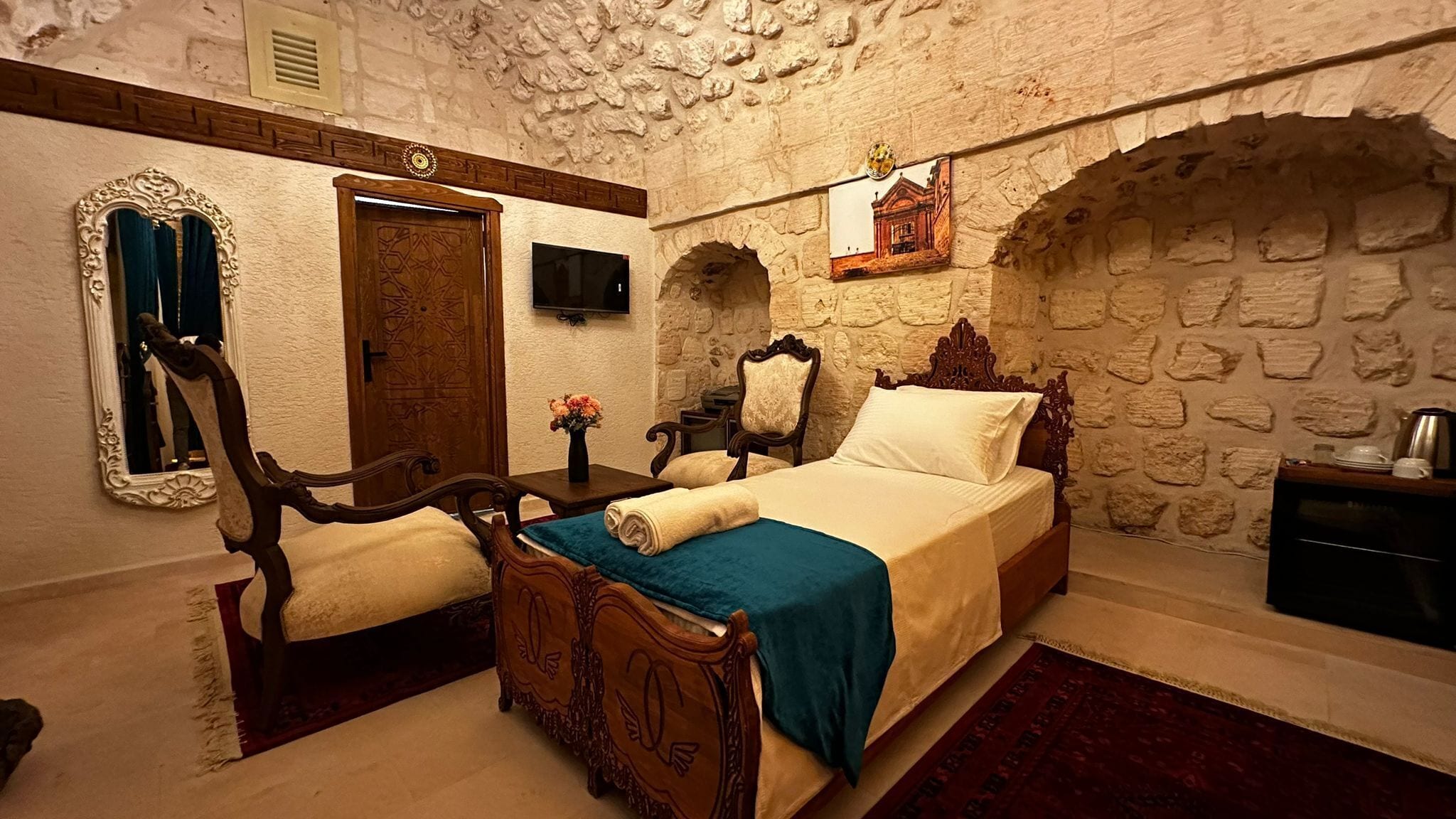 Una habitación con pared de piedra, una cama con colcha azul, una silla de madera, un escritorio de madera y un espejo con marco ornamentado.