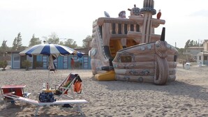 Ein Strand mit einer großen, gestreiften, schiffartigen Konstruktion, einem Strandkorb und einer Liege mit einer Tasche.
