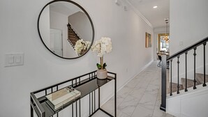 A hallway with a round mirror, a console table with a vase of white flowers, a staircase with black railings, and a dining area in the background.