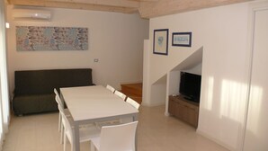 Een moderne woonkamer met een witte eettafel, witte stoelen, een donkere bank, een flatscreen televisie op een houten dressoir en een trap naar een ander niveau.