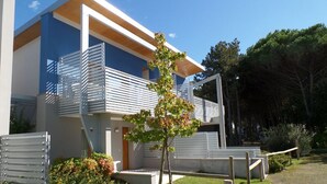 Un edificio moderno de dos pisos con un balcón, un pequeño árbol y una zona pavimentada con un banco.