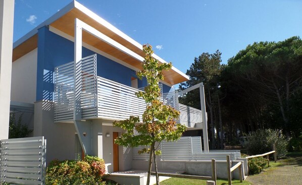 A modern two-story building with a balcony, a small tree, and a paved area with a bench.