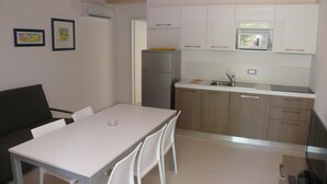 Una cocina compacta con armarios blancos, un microondas integrado y un refrigerador. Hay una mesa de comedor con sillas y un sofá en la zona de estar contigua.