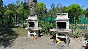 Dos hornos de ladrillo al aire libre con un área pavimentada en frente, rodeados de follaje y una cerca verde.